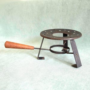 Peut inclure: Un support de pot à fondue en métal vintage avec un dessus circulaire et une poignée en bois. Le support est en métal foncé et possède un anneau pour maintenir le pot. La poignée en bois est marron clair.