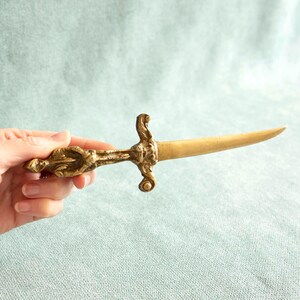 May include: A vintage letter opener with a brass finish. The handle features an ornate, sculpted design, and the blade is slender and pointed. The overall aesthetic suggests a historical or antique style, suitable for a desk or office setting.