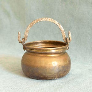 May include: A small, antique brass pot with a curved handle. The pot has a rounded body and a textured surface, with decorative detailing on the handle and the points where it attaches to the pot.