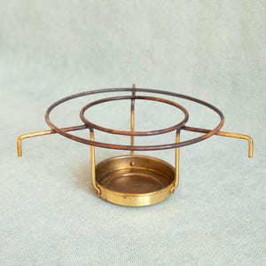 May include: A vintage brass and metal candle holder. The candle holder has a circular base and a series of concentric rings above it. The metal has a warm, aged patina. Two curved handles extend from the sides.