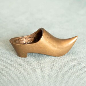 May include: A small, bronze-colored decorative clog. The miniature shoe has a pointed toe and a small heel. The interior of the clog is hollow. The clog is sitting on a light blue surface.