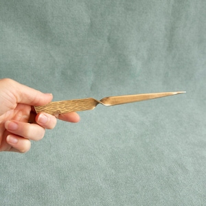 May include: A brass letter opener with a textured handle and a pointed blade. The handle has a wood grain pattern. The letter opener is held against a muted green background.