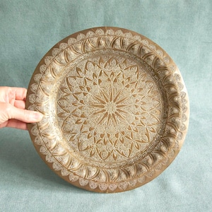 Può includere: Un vassoio rotondo in ottone antico con intricati motivi floreali e geometrici. Il vassoio ha un bordo rialzato e un design centrale dettagliato, che mostra un'estetica vintage. Il vassoio è tenuto da una mano.