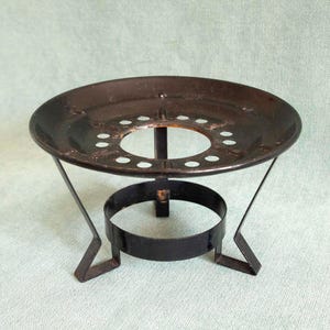 May include: A vintage black metal trivet. The round top has a central hole and smaller holes. It has a circular base and three angled legs. Designed to elevate a pot or dish, it measures approximately 15 cm in diameter.