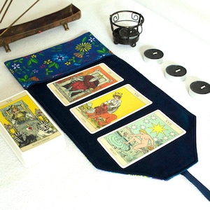 May include: A navy blue tarot card reading mat with a floral patterned pocket. Three tarot cards are displayed on the mat, with a deck of cards to the left. An incense burner, candles, and a decorative candle holder are also present.