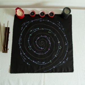 Puede incluir: Un paño de tela negro cuadrado con un diseño de espiral central bordado con hilo azul claro y pequeños acentos coloridos. El paño está rodeado por un portavelas rojo, tres pequeños portavelas de té rojos, una vela negra y un porta incienso.