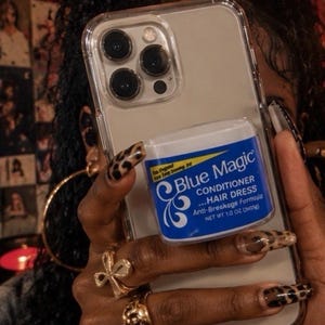 May include: A smartphone with a clear case featuring a Blue Magic Conditioner Hair Dress Anti-Breakage Formula container attached to the back. The label is blue and white with yellow accents, clearly stating the product name and net weight 1/2 oz.