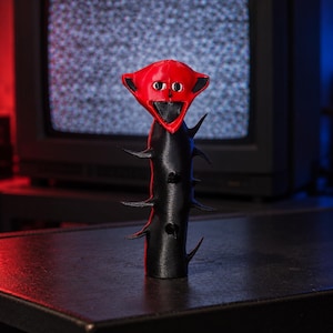 May include: A 3D printed figurine featuring a black cylindrical base with protruding spikes and a red head with a wide, open mouth, black eyes, and black ear-like appendages. The figure has a smooth, matte finish and stands upright.