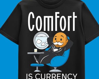 May include: Black t-shirt with the words "Comfort Is Currency" in white. The shirt features a cartoon image of a man in a tuxedo relaxing in a blue chair. A small table with a drink and a fishbowl are also visible.