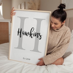 Könnte beinhalten: Ein gerahmter Druck mit einem hellen Holzrahmen. Der Druck zeigt einen großen grauen Buchstaben "H" mit dem Namen "Hawkins" in schwarzer Schrift. Unter dem Namen steht der Text "Est. 2019" in kleinerer Schrift. Der Hintergrund ist weiß.