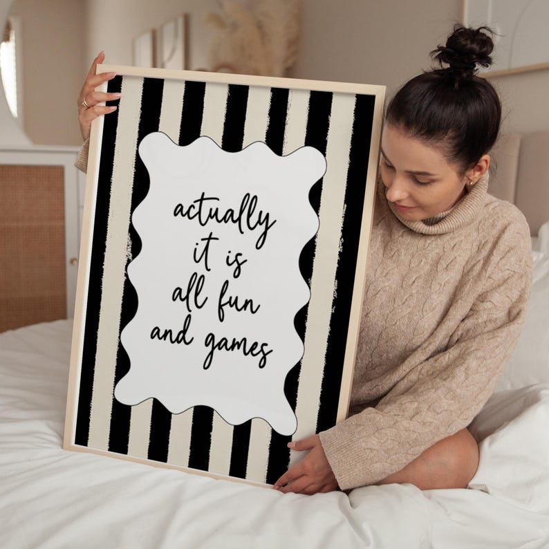 May include: A framed art print with a black and beige striped background. The print features a white, wavy-edged shape with the words "actually it is all fun and games" in black script. The frame is a light wood color.