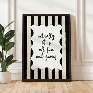 May include: Black framed wall art with a black and white striped background. The art features a white, wavy-edged shape with the words "actually it is all fun and games" in black script. The art is displayed on a wooden floor.