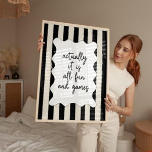 May include: A framed art print with a black and white striped background. The print features a white, wavy-edged shape with the words "actually it is all fun and games" in black script. The frame is a light wood color.