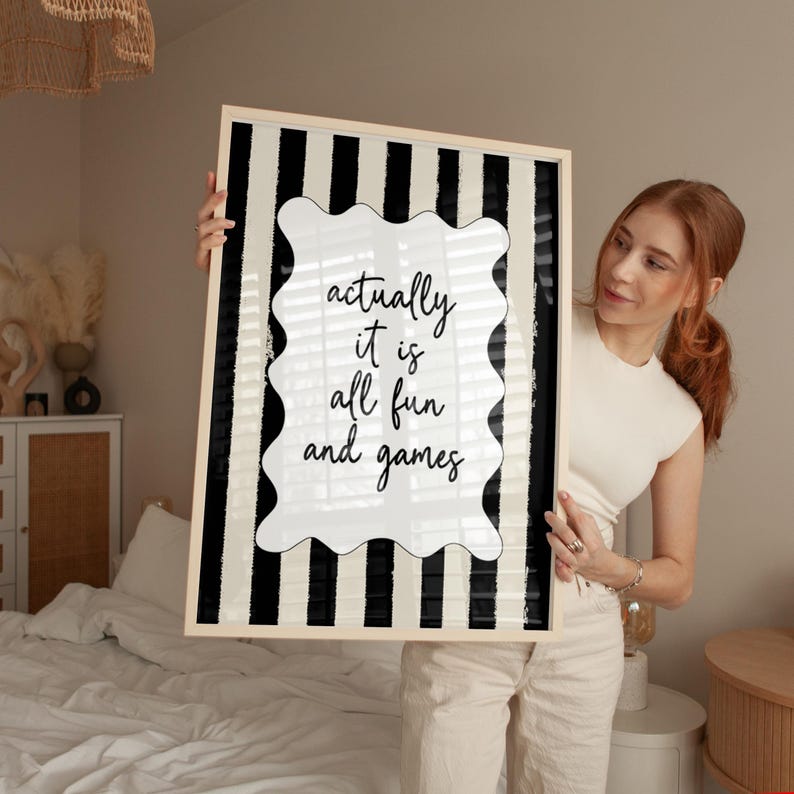 May include: A framed art print with a black and white striped background. The print features a white, wavy-edged shape with the words "actually it is all fun and games" in black script. The frame is a light wood color.