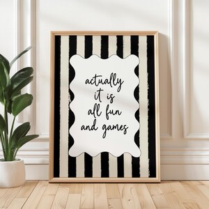 May include: A framed print with a black and cream striped background. The print features a white, wavy-edged shape with the words "actually it is all fun and games" in black script. The frame is a light wood color.