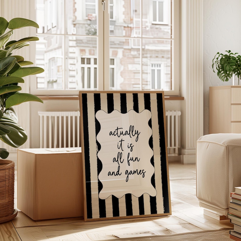 May include: A framed art print with a black and white striped border. The print features the phrase "actually it is all fun and games" in a decorative, wavy-edged frame. The artwork is leaning against a box on a wooden floor.