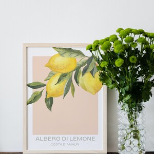 Op de afbeelding: Een ingelijste print van een aquarel van een tak van een citroenboom met drie citroenen. De print heeft een perzikkleurige achtergrond en de tekst "ALBERO DI LEMONE COSTA D'AMALFI" in grijze letters. De print ligt op een houten tafel naast een transparante glazen vaas met groene bloemen.