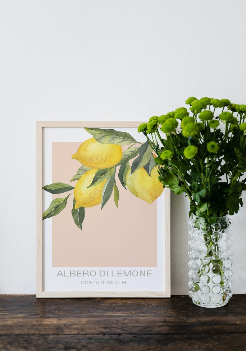 Op de afbeelding: Een ingelijste print van een aquarel van een tak van een citroenboom met drie citroenen. De print heeft een perzikkleurige achtergrond en de tekst "ALBERO DI LEMONE COSTA D'AMALFI" in grijze letters. De print ligt op een houten tafel naast een transparante glazen vaas met groene bloemen.