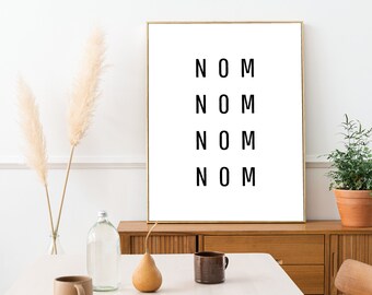 Op de afbeelding: Een ingelijste kunstafdruk met het woord "NOM" vier keer in zwart op een witte achtergrond. De lijst is goudkleurig. De afbeelding toont ook een houten kast, een plant in een terracotta pot en decoratieve items.