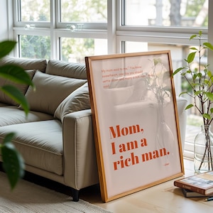 Könnte beinhalten: Ein gerahmter Druck mit dem Text "Mom, I am a rich man" in orangefarbenen Buchstaben. Der Druck lehnt an einem beigen Sofa in einem hellen Raum. Eine Glasvase mit Grünzeug befindet sich neben dem Druck.