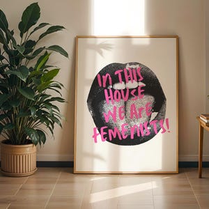 Könnte beinhalten: Gerahmter Kunstdruck mit einem schwarz-weißen Lippenmotiv und dem pinkfarbenen Text "IN THIS HOUSE WE ARE FEMINISTS!". Das Kunstwerk wird an einer Wand neben einer Topfpflanze präsentiert.
