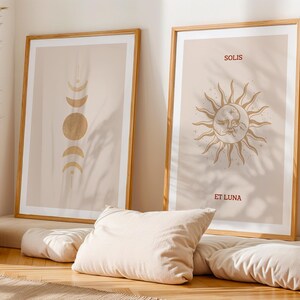 Könnte beinhalten: Zwei gerahmte Kunstdrucke. Einer zeigt goldene Mondphasen, der andere eine Sonne mit Gesicht und den Worten "SOLIS ET LUNA" in Burgunderrot. Beide sind in Holzrahmen und lehnen an einer Wand.