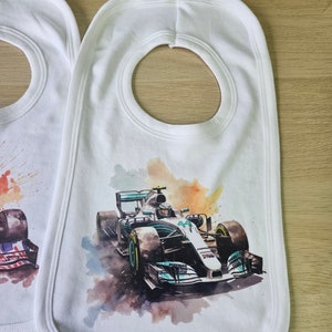 May include: Two white baby bibs with a watercolour design of a Formula 1 racing car. The bib on the right has a green and white racing car with the number 44 on it. The bib on the left has a red, white and blue design.