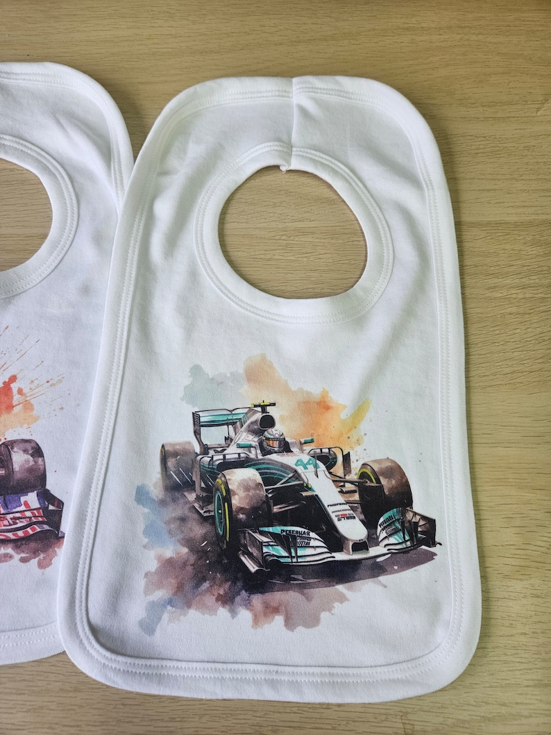 May include: Two white baby bibs with a watercolour design of a Formula 1 racing car. The bib on the right has a green and white racing car with the number 44 on it. The bib on the left has a red, white and blue design.