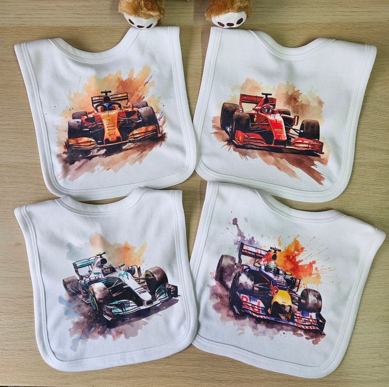 May include: Four white bibs with watercolour images of Formula One racing cars. The bibs feature different racing cars, including a red Ferrari and a blue Red Bull.