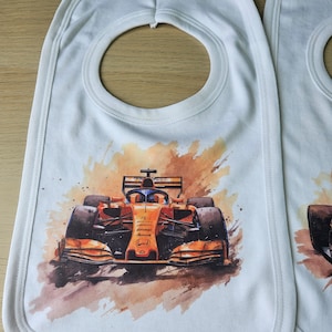 May include: White baby bib featuring a watercolour image of a Formula 1 racing car. The car is orange and black with the Honda logo on the side. The bib has a white trim around the edges.