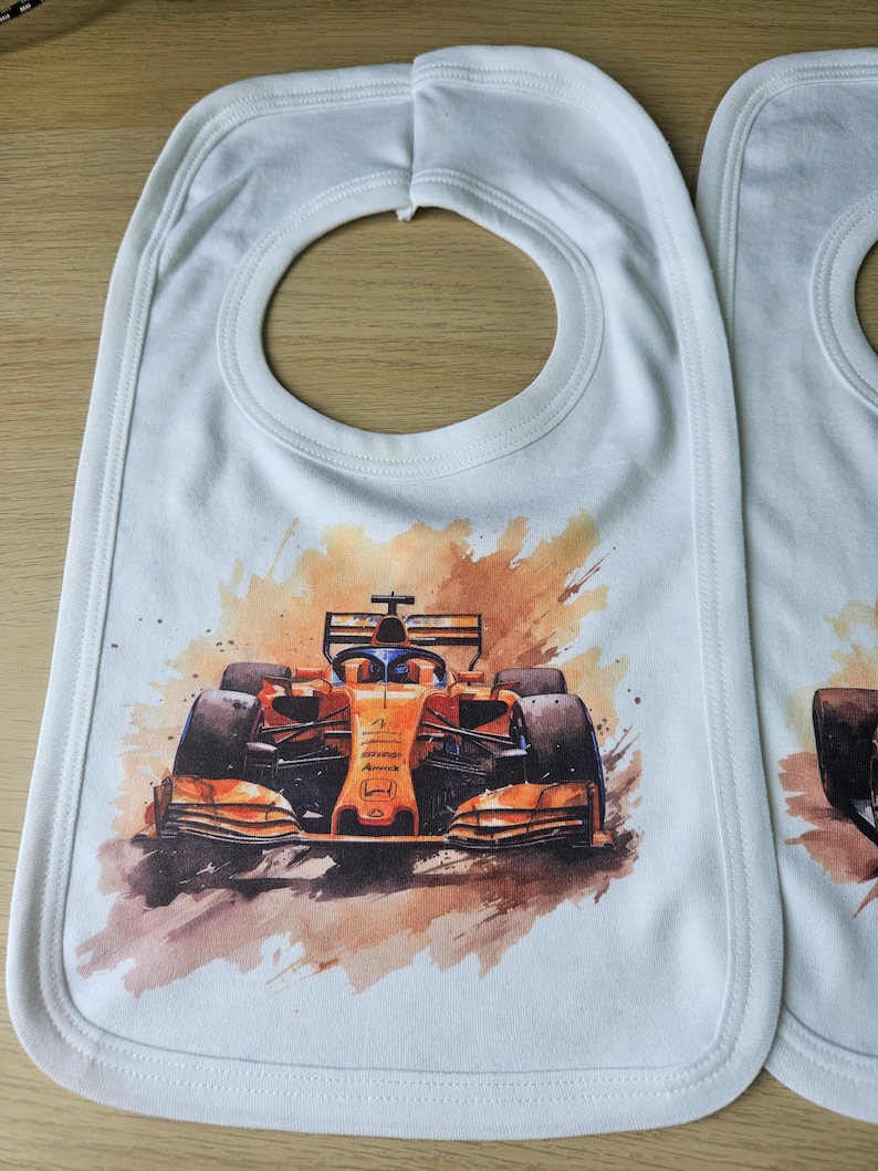 May include: White baby bib featuring a watercolour image of a Formula 1 racing car. The car is orange and black with the Honda logo on the side. The bib has a white trim around the edges.