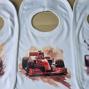 May include: Three white bibs with a red race car printed on the centre bib. The race car is a watercolour illustration with a brown background.