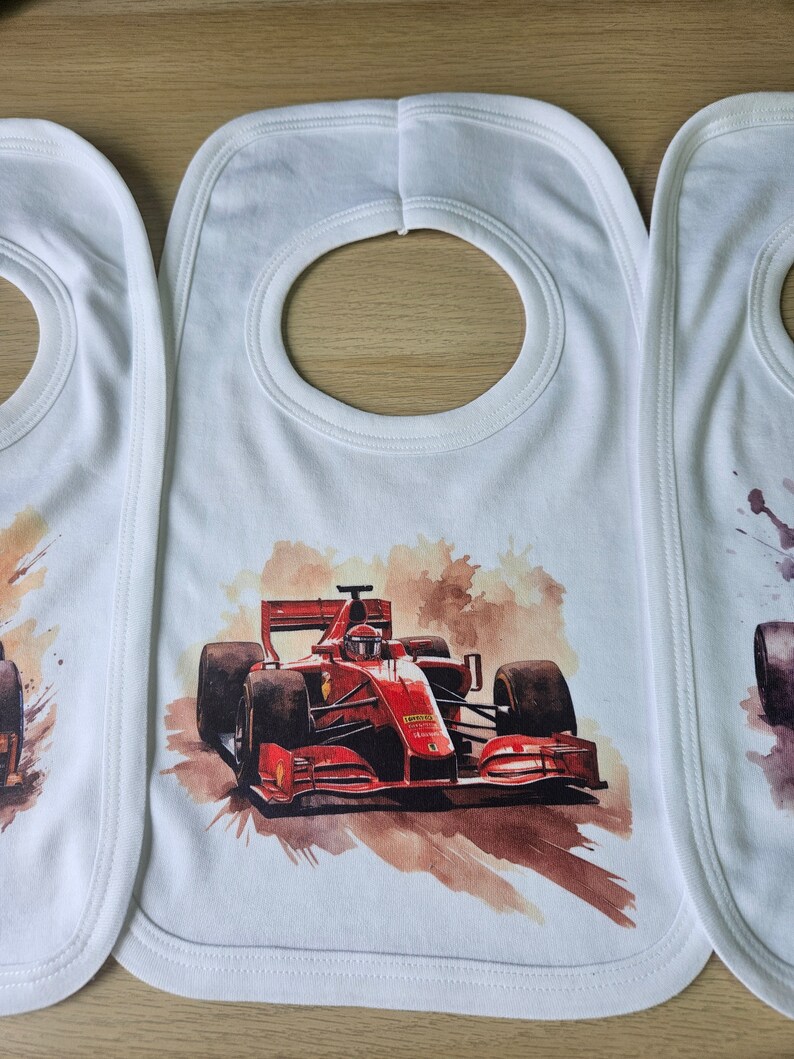 May include: Three white bibs with a red race car printed on the centre bib. The race car is a watercolour illustration with a brown background.