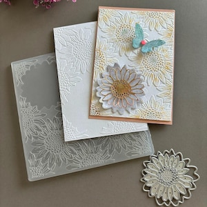 May include: Handmade paper crafts with sunflower designs. Includes a white embossed card with a 3D sunflower and a blue butterfly, a clear embossing folder, and a metal sunflower die cut.