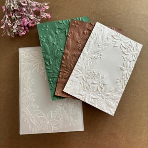 May include: A collection of embossed paper cards in various colors: white, green, and brown. The cards feature floral and leaf designs, with a clear plastic embossing folder in the foreground. Decorative pink flowers are in the upper left corner.
