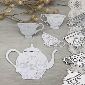 Op de afbeelding: Een verzameling witte metalen stansvormen met een theepot en verschillende theekopjes. Elk stuk vertoont ingewikkelde reliëfpatronen, waaronder geschulpte randen, bloemmotieven en lineaire details, ontworpen voor knutsel- en papierprojecten.