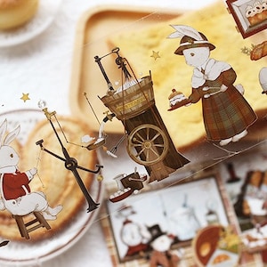 Op de afbeelding: Een vel met transparante stickers met illustraties van konijnen, gebak en vintage bakkerijapparatuur. De stickers zijn bedrukt met de tekst "Workshop -" en "Choux a la Creme".