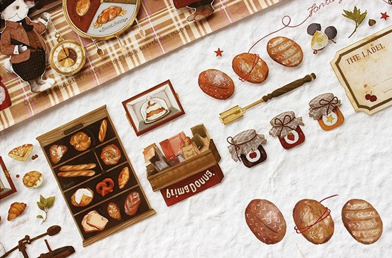 Op de afbeelding: Een verzameling ge&iuml;llustreerde stickers met verschillende bakkerijproducten, waaronder brood, gebak en potten jam. De stickers zijn op een witte achtergrond met een geruit patroon in de linkerbovenhoek. De tekst "Little Downs" is zichtbaar op een van de stickers.