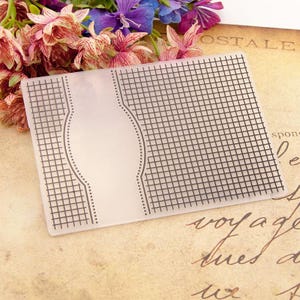 May include: A clear plastic craft stencil with a grid pattern and a large oval shape in the center. The stencil is designed for use with crafting projects.