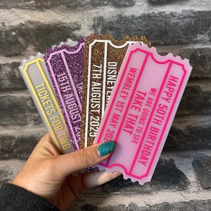 May include: Four pink, purple, gold, and white glitter ticket-shaped cards with text that reads "Happy 50th Birthday", "We are going to see Take That", "Wembley 1st May 202", "Disney", "7th August 2023", "15th August", and "Tickets for Two".