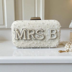 Pode incluir: Uma clutch retangular branca coberta por pequenas p&eacute;rolas. O texto "MRS B" est&aacute; escrito em letras prateadas e strass. A clutch tem um fecho de madeira e est&aacute; colocada numa superf&iacute;cie branca.