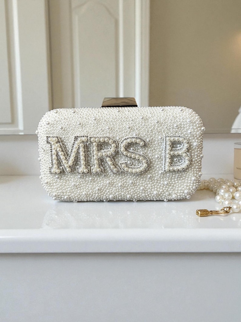 Pode incluir: Uma clutch retangular branca coberta por pequenas p&eacute;rolas. O texto "MRS B" est&aacute; escrito em letras prateadas e strass. A clutch tem um fecho de madeira e est&aacute; colocada numa superf&iacute;cie branca.