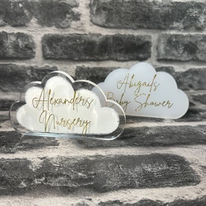 May include: Two white acrylic cloud-shaped signs with gold lettering. One sign reads "Alexander's Nursery" and the other sign reads "Abigail's Baby Shower".