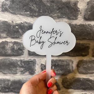 May include: A white cloud-shaped sign with the text "Jennifer's Baby Shower" in elegant script. The sign is held in front of a grey brick wall. The sign is designed for a baby shower celebration.