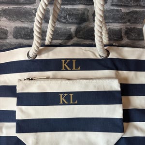 May include: A large, striped tote bag with rope handles. The bag is white and navy blue with the initials "KL" embroidered on the front. A smaller matching zippered pouch is included with the same initials.
