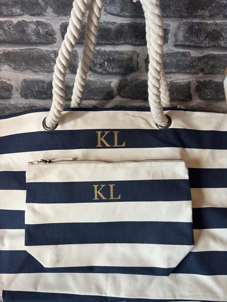 May include: A large, striped tote bag with rope handles. The bag is white and navy blue with the initials "KL" embroidered on the front. A smaller matching zippered pouch is included with the same initials.