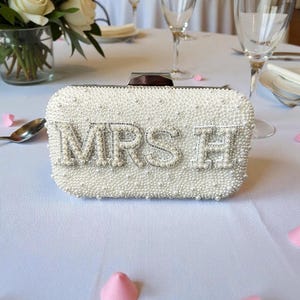 Pode incluir: Uma clutch branca com contas com as letras "MRS H" em prata. A bolsa retangular &eacute; coberta por pequenas p&eacute;rolas e tem um fecho prateado. A clutch est&aacute; sobre uma toalha de mesa branca, com p&eacute;talas rosa espalhadas.