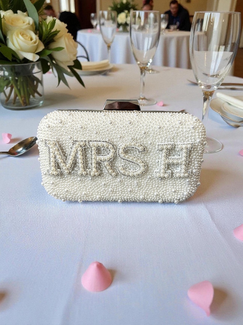 Pode incluir: Uma clutch branca com contas com as letras "MRS H" em prata. A bolsa retangular &eacute; coberta por pequenas p&eacute;rolas e tem um fecho prateado. A clutch est&aacute; sobre uma toalha de mesa branca, com p&eacute;talas rosa espalhadas.