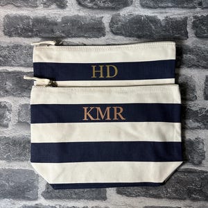 May include: Two striped canvas zippered pouches with gold lettering. The top pouch has the initials "HD" and the bottom pouch has the initials "KMR".