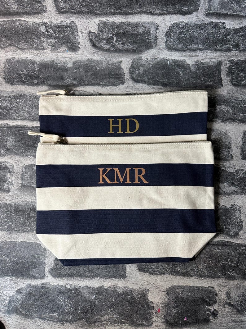 May include: Two striped canvas zippered pouches with gold lettering. The top pouch has the initials "HD" and the bottom pouch has the initials "KMR".
